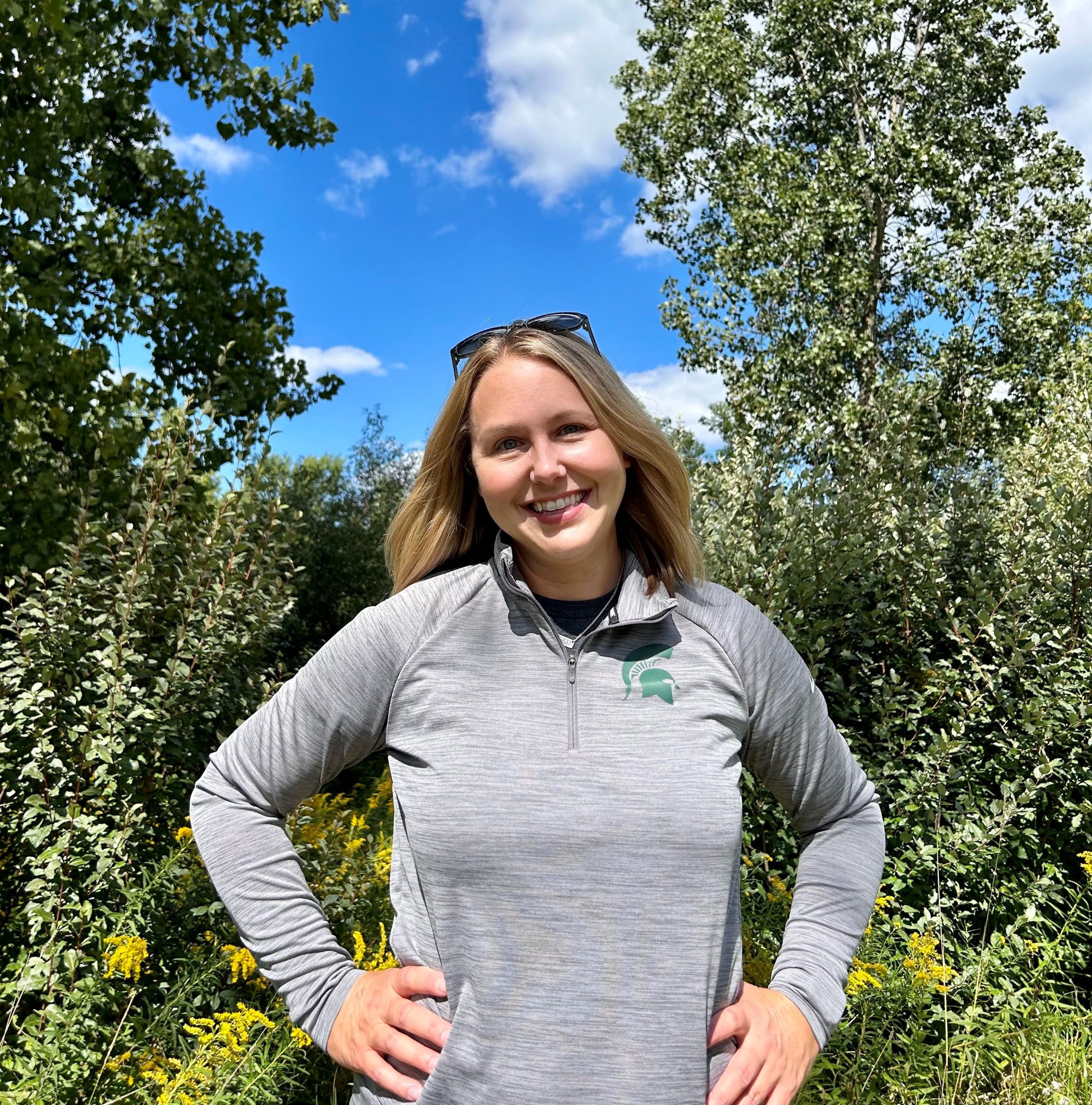 Outdoor portrait of Jennifer Petzko standing with hands on hips in front of green trees and shrubs under a blue sky.
